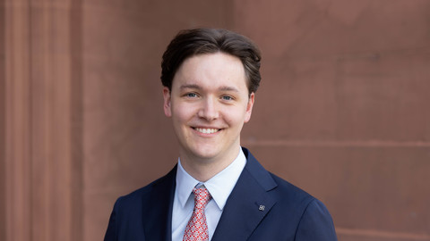 Henrik Sauer. He is wearing a dark blue suit with a salmon-colored tie and is standing in front of a brown wall.