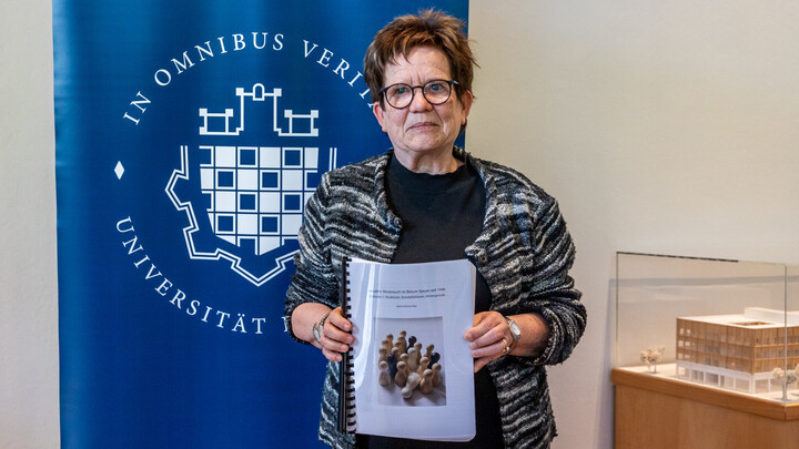 Prof. Dr. Sylvia Schraut steht vor dem Uni Mannheim Logo und hält einen Ausdruck ihrer Studie in den Händen