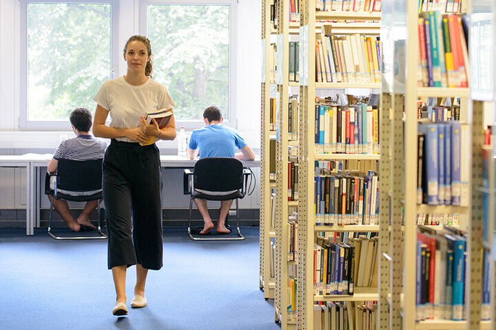 Eine junge Frau hält ein paar Bücher in ihrem Arm und geht durch eine Bibliothek. Im Hintergrund lernen Studenten an Einzeltischen.