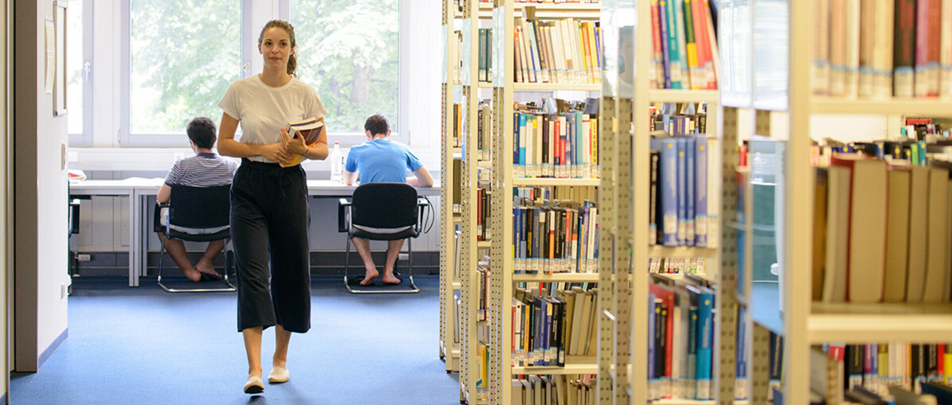 Eine junge Frau hält ein paar Bücher in ihrem Arm und geht durch eine Bibliothek. Im Hintergrund lernen Studenten an Einzeltischen.