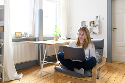 Eine junge Frau sitzt in einem gemütlichen Zimmer mit ihrem Laptop auf einem Sessel.