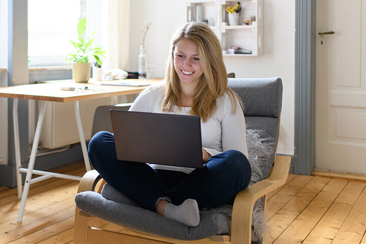 Eine junge Frau sitzt in einem gemütlichen Zimmer mit ihrem Laptop auf einem Sessel.