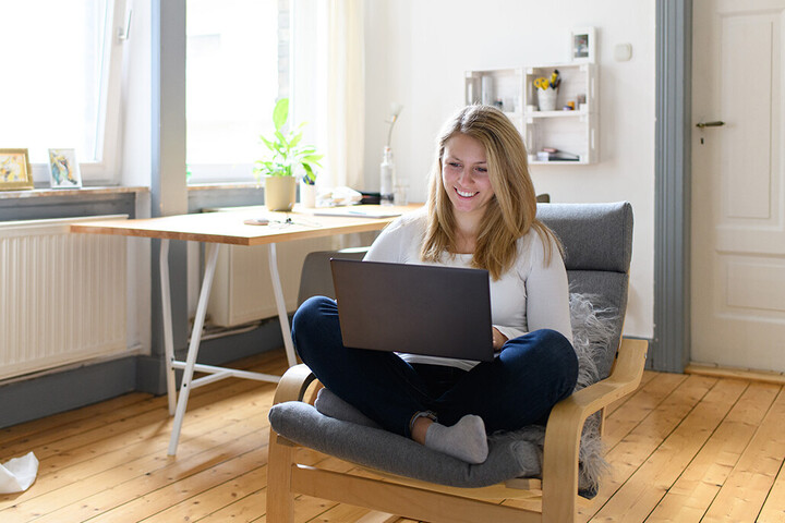 Eine junge Frau sitzt in einem gemütlichen Zimmer mit ihrem Laptop auf einem Sessel.
