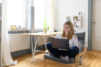 Eine junge Frau sitzt in einem gemütlichen Zimmer mit ihrem Laptop auf einem Sessel.