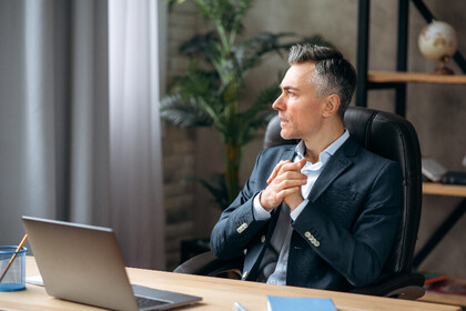 A man in a suit is sitting at a desk. There is an open laptop on the desk. The man looks thoughtfully to the side.