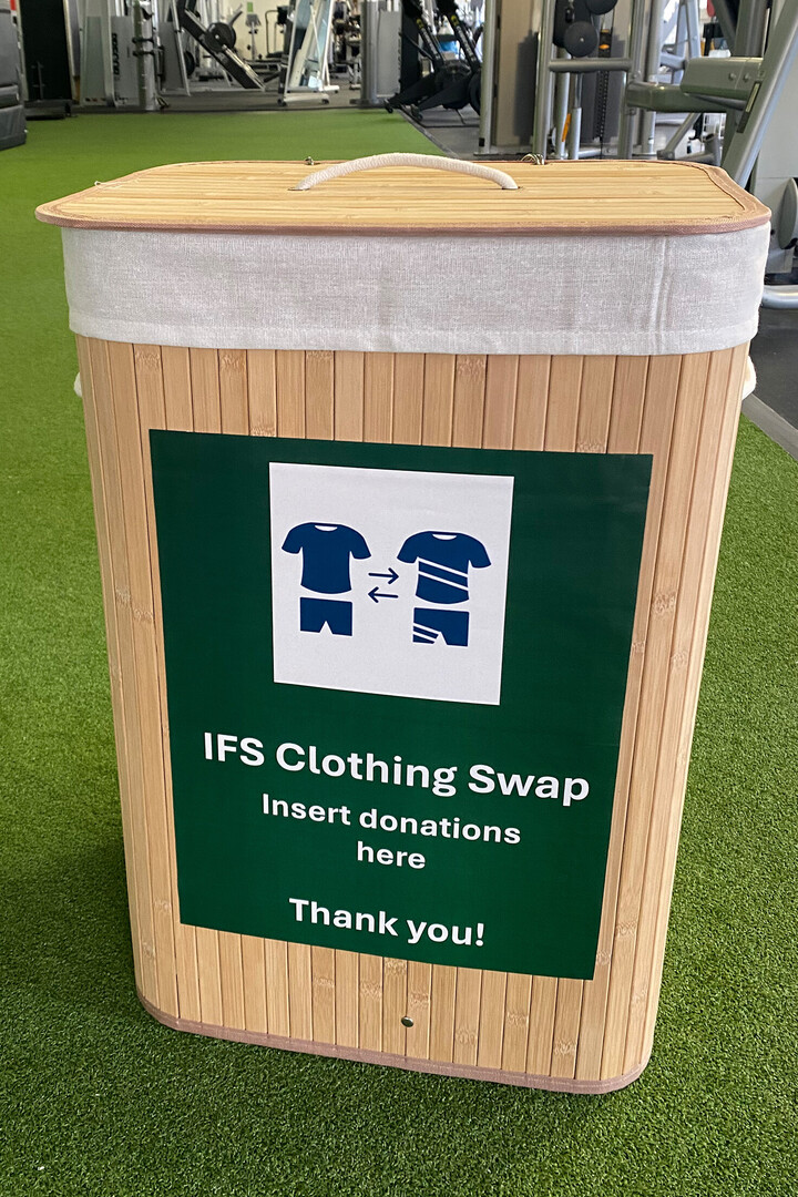 Eine helle Holzbox steht auf dem grünen Teppich in einem Fitnessstudio. Auf der Box steht "IFS Clothing Swap". Im Hintergrund sind Fitnessgeräte zu sehen.