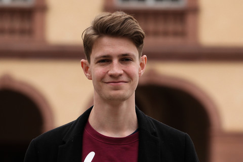 Jan Bleil hat kurze, hellbraune Haar. Er trägt ein weinrotes Shirt mit weißer Aufschrift, darüber eine lockeren schwarze Jacke. 