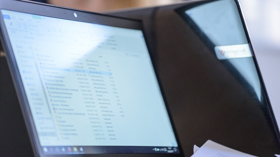 A screen displaying a list of files is illuminated by the sun. Next to it  is another laptop, which can be seen from behind.