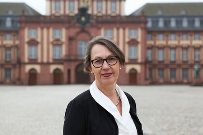 Portrait von Sabine Sonnentag. Sie trägt eine weiße Bluse sowie einen schwarzen Blazer und steht auf dem Ehrehof vor dem Schloss.  