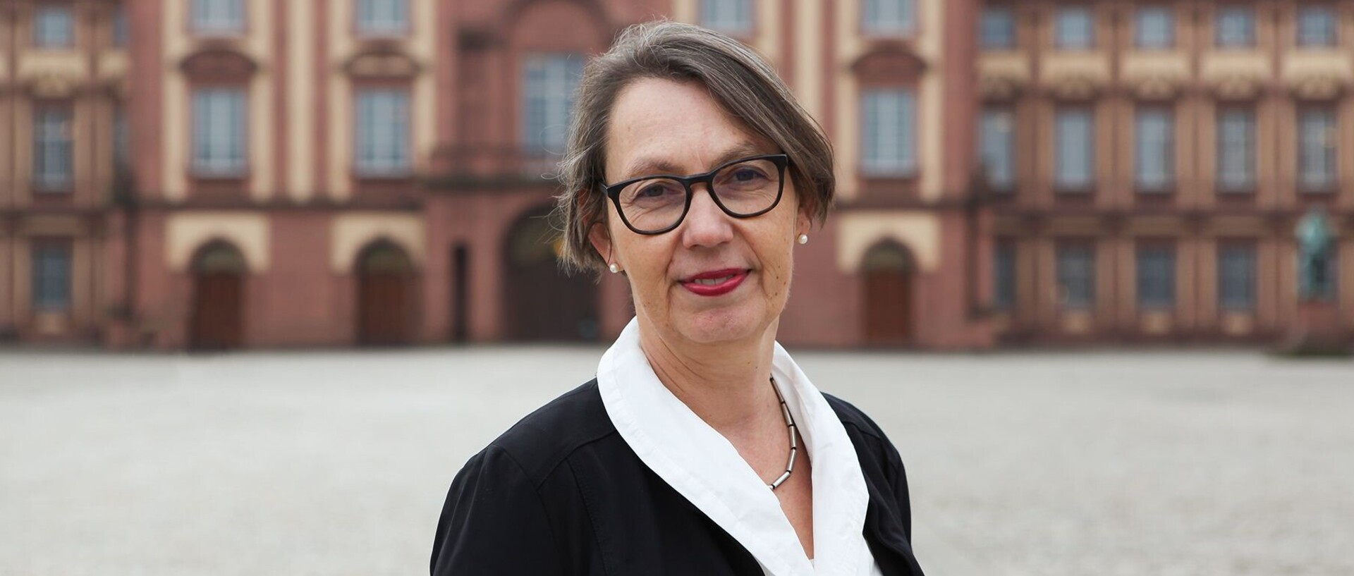 Portrait von Sabine Sonnentag. Sie trägt eine weiße Bluse sowie einen schwarzen Blazer und steht auf dem Ehrehof vor dem Schloss.  