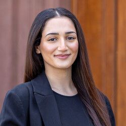 A portrait of the student Helin Yalman. She is wearing a black top with a black blazer. Her long brown hair is worn loose with a center part. Helin Yalman is looking directly at the camera and smiling.