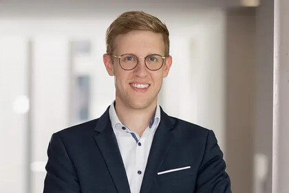 Portrait von Jens Förderer im Anzug. Er steht lächelnd in einem hellen Flur.