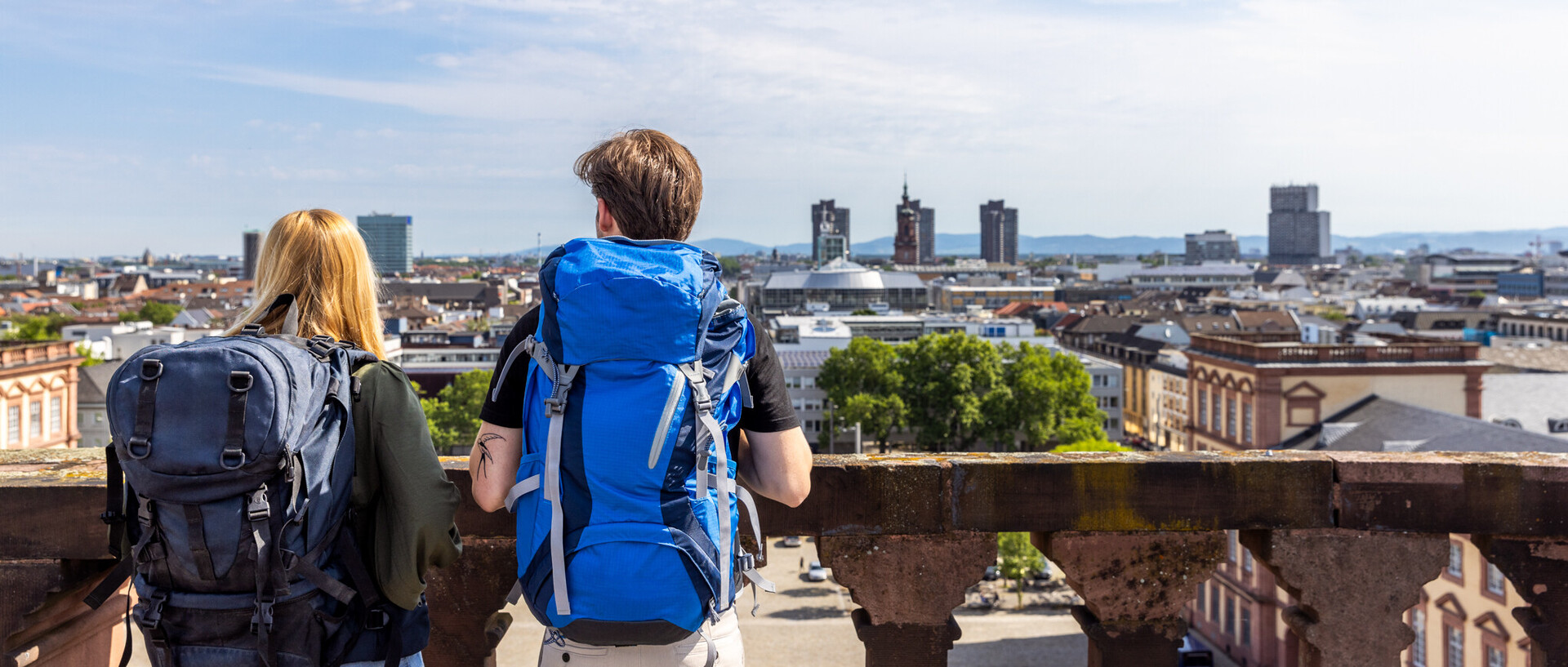 Zwei Studierende mit Reiserucksäcken auf dem Schlossdach Mannheims. Sie haben einen Blick auf den Ehrenhof und die Stadt.