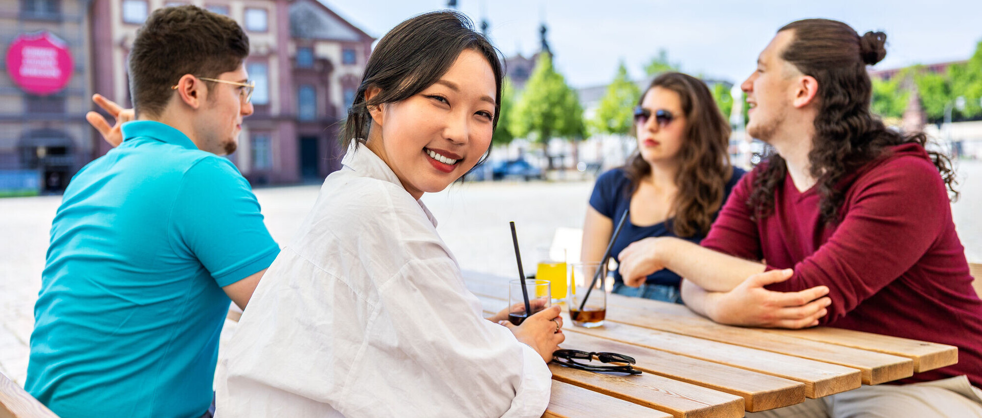 Vier Studierende sitzen an einem Café-Tisch. Eine Person schaut in die Kamera. Im Hintergrund ist ein Teil des Schlosses zu sehen.