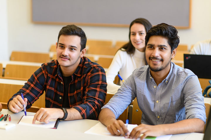 Zwei Studenten sitzen an einem Tisch in einem Seminarraum. Vor ihnen liegen Papier und Stifte.