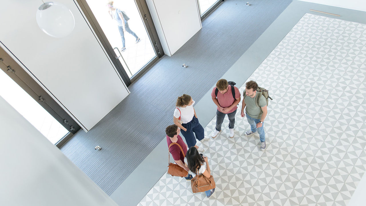 Zwei Studentinnen und drei Studenten unterhalten sich in dem hellen Eingangsbereich des Gebäudes B6 der Universität Mannheim. Sie wurden von einem höheren Stockwerk aus fotografiert.