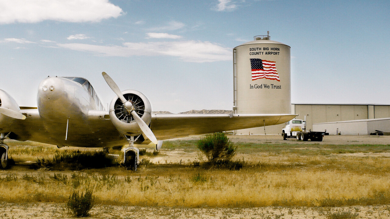 Im Vordergrund steht ein Flugzeug, dahinter ein Silo mit der USA-Flagge und der Aufschrift "South Big Horn County Airport, In God We Trust".