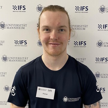 Florian Haupt hat seine langen braunen Haare zu einem Zopf gebunden. Er trägt ein schwarzes T-Shirt mit dem Logo des Instituts für Sport.