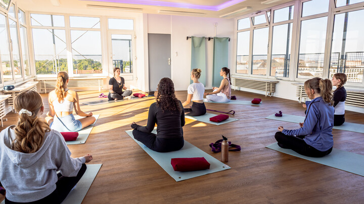 Bild aus dem SkyStudio N6. Hinten sitzt Charlotte Werner auf einer Yoga-Matte, mit dem Rücken zur Kamera mehrere Teilnehmende im Schneidersitz.