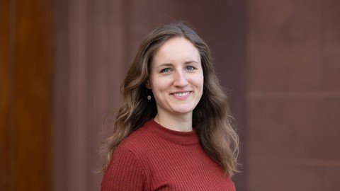 Milena Rapp. She is wearing a red sweater and is standing in front of brown stonewall.