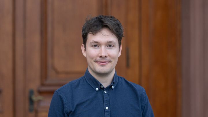 Maik Sattelmaier. He is wearing a blue shirt and is standing in front of one of the castle doors.