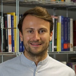 Eine Person trägt ein graues Hemd und steht vor einem Bücherregal. Die Person heißt Maximilian Beicher.