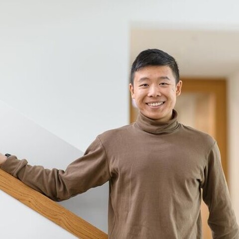 Jianming Cui aus Kanton. Er trägt einen braunen Rollkragenpullover und legt seinen rechten Arm lässig auf ein Treppengeländer.