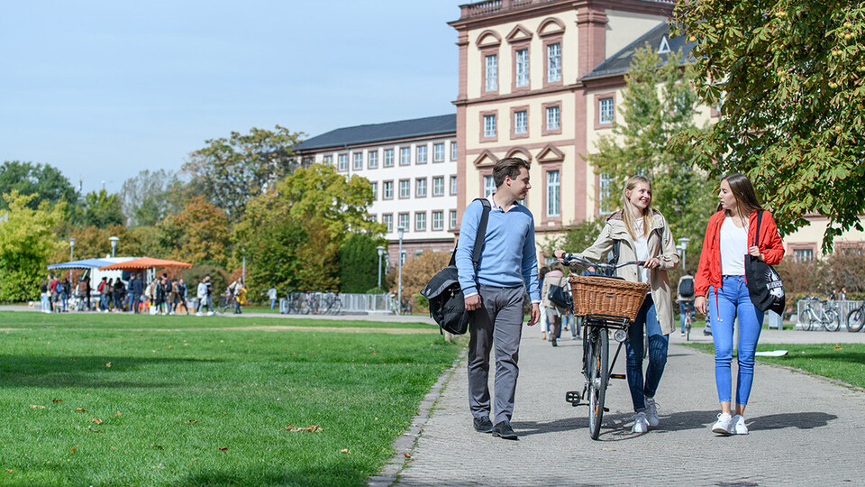 Drei junge Menschen unterhalten sich auf einem Gehweg vor dem Mannheimer Schloss im Mensapark, die Person in der Mitte schiebt ein Fahrrad.