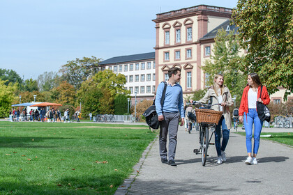 Drei junge Menschen unterhalten sich auf einem Gehweg vor dem Mannheimer Schloss im Mensapark, die Person in der Mitte schiebt ein Fahrrad.