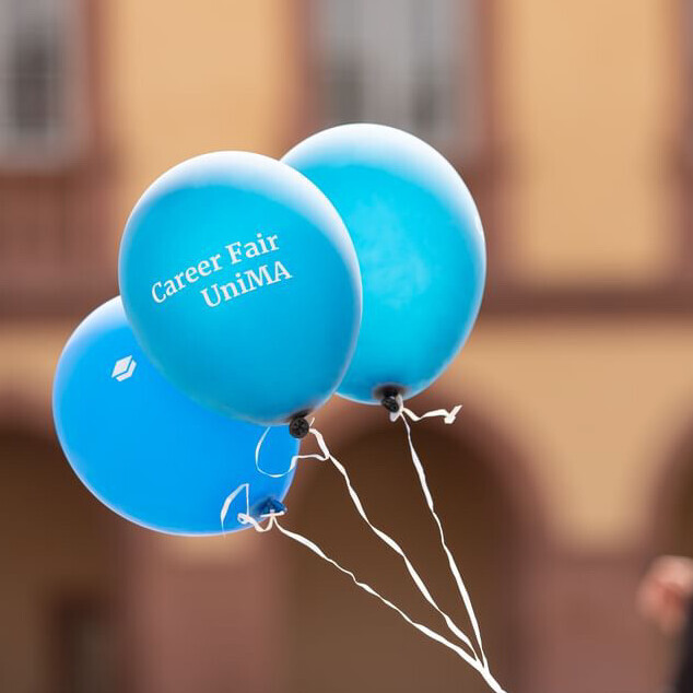 Drei blaue Luftballons, auf einem steht Carer Fair. Im Hintergrund sieht man verschwommen zwei Personen und das Schloss.