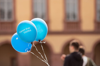 Drei blaue Luftballons, auf einem steht Carer Fair. Im Hintergrund sieht man verschwommen zwei Personen und das Schloss.
