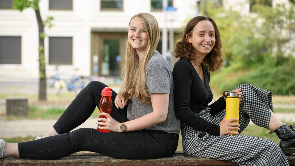 Zwei Studentinnen lehnen Rücken an Rücken draußen auf einer Bank. Sie haben beide eine Trinkflasche in der Hand.