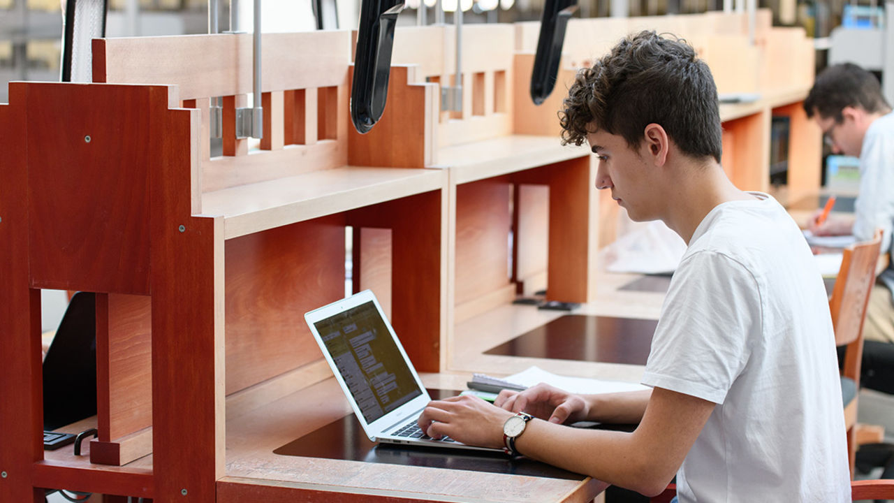 Ein Student arbeitet im Bibliotheksbereich A3 am Laptop. Er trägt ein weißes T-Shirt und sitzt an einem langen Holztisch.