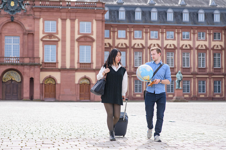 A woman with a bag and a suitcase and a man who is holding a globe are walking on the Ehrenhof in front of the castle.