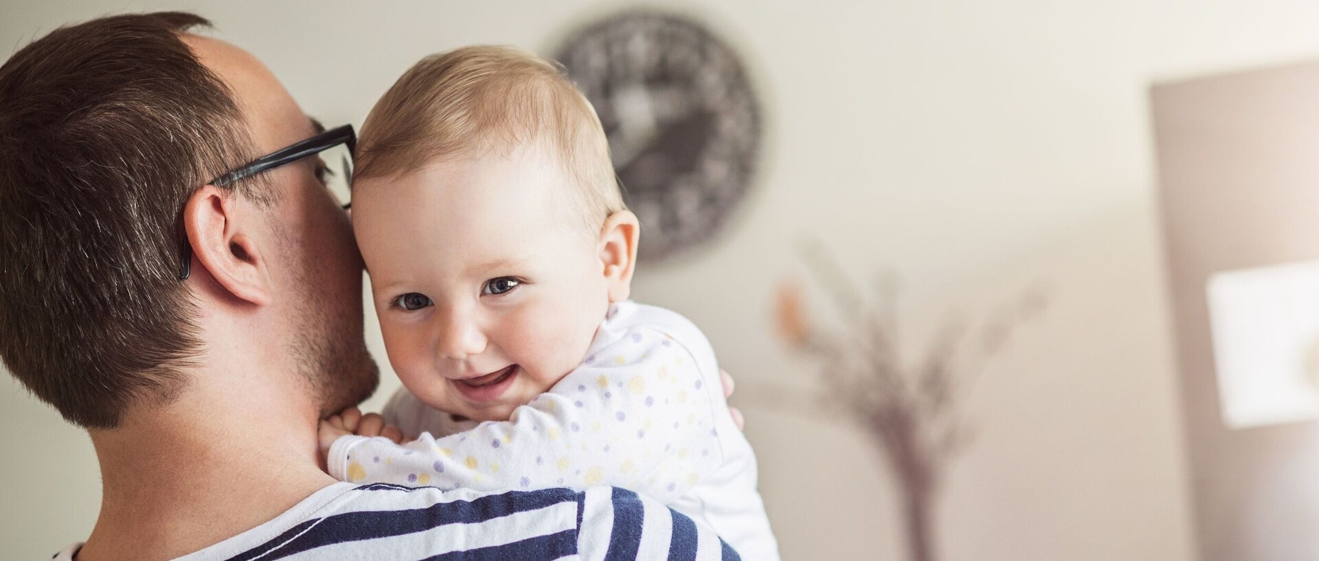 Mann mit blau weiß gestreiften T-Shirt hat Baby auf dem Arm. Baby lächelt in die Kamera und hat einen weißen Body mit gelb-blauen Punkten an.