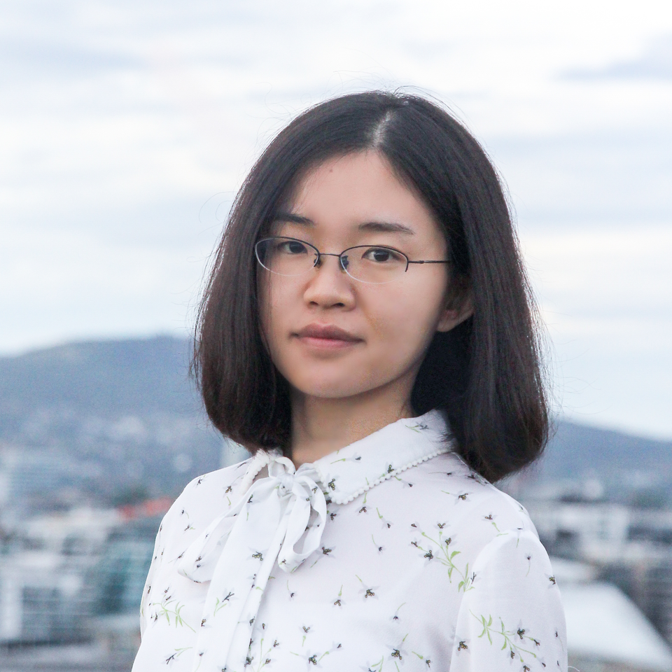 Lei Li im Freien, im Hintergrund sieht man einen Berg und eine Stadt. Sie trägt eine Brille und eine geblümte Bluse mit Schleife.