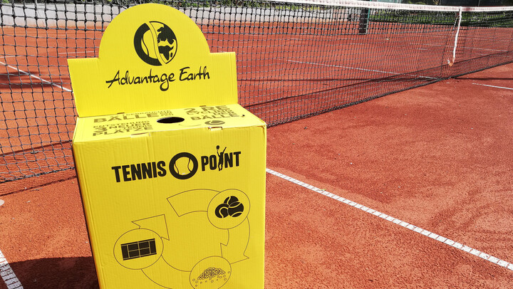 Eine gelbe Pappbox steht auf einem Tennisfeld. Auf der Box steht "Advantage Earth" und "Tennis Point". Hinter der Box ist das Tennisnetz aufgespannt.
