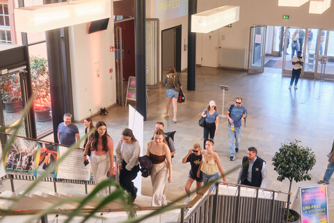 Personen steigen die Treppe im Foyer des Ostflügels der Uni Mannheim hoch, im Hintergrund sind Plakate und eine Projektion der Veranstaltung "Uni-Fest".