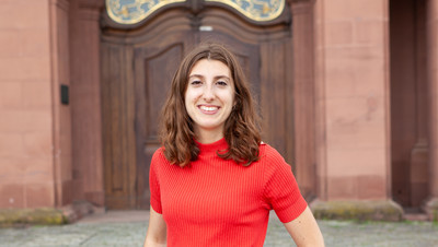 Eine lächelnde Person trägt ein rotes T-Shirt und steht vor einer braunen Schlosstüre. Die Person heißt Léa Beringuer.