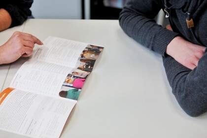 A brochure lies on the table. Two people, only their arms are visible, are looking at it.