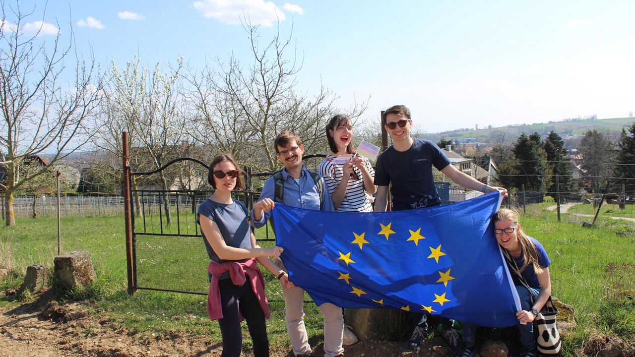 Drei Studentinnen und zwei Studenten halten eine Flagge der Europäischen Union hoch. Sie sind sommerlich gekleidet. Im Hintergrund stehen viele Bäume.