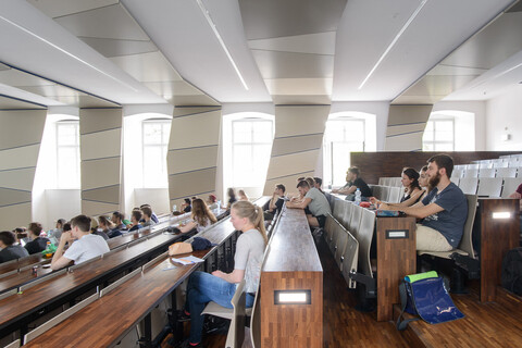 Studierende in einem hölzernen, lichtdurchfluteten Hörsaal mit vielen Fenstern