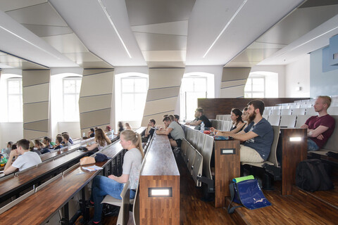 Studierende in einem hölzernen, lichtdurchfluteten Hörsaal mit vielen Fenstern