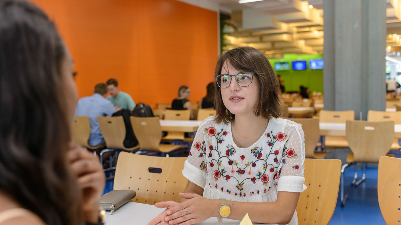 Zwei Studentinnen sitzen gemeinsam an einem Tisch in der Mensa und unterhalten sich.