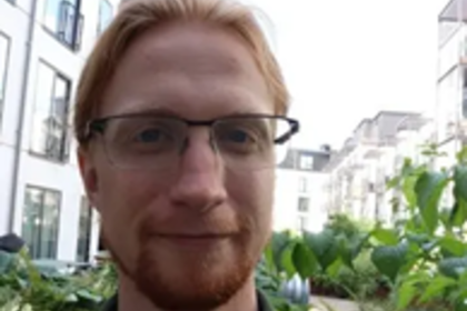 Foto von John Collins im Freien. Er hat lange rotblonde Haare und trägt eine Brille und ein grünes Hemd.