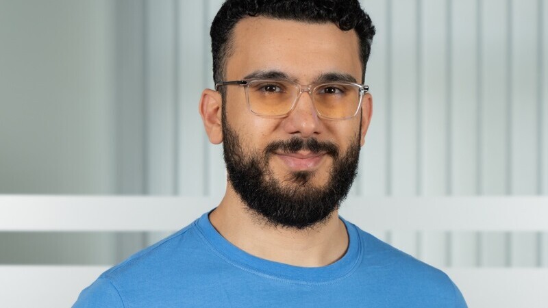 Porträt von Nader Hotait. Er hat kurze schwarze Haare, einen Bart und eine Brille. Er trägt ein blaues T-Shirt.