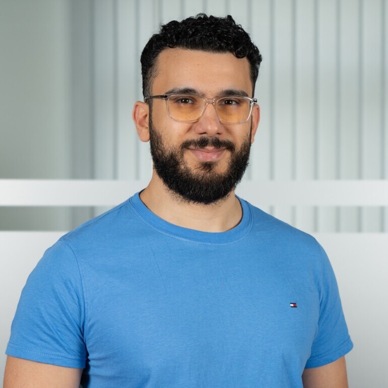 Porträt von Nader Hotait. Er hat kurze schwarze Haare, einen Bart und eine Brille. Er trägt ein blaues T-Shirt.