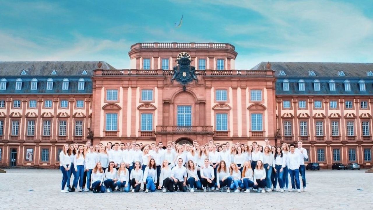 Studierende posieren vor dem Mannheimer Schloss. Sie tragen alle ein weißes Oberteil und eine blaue Jeans.