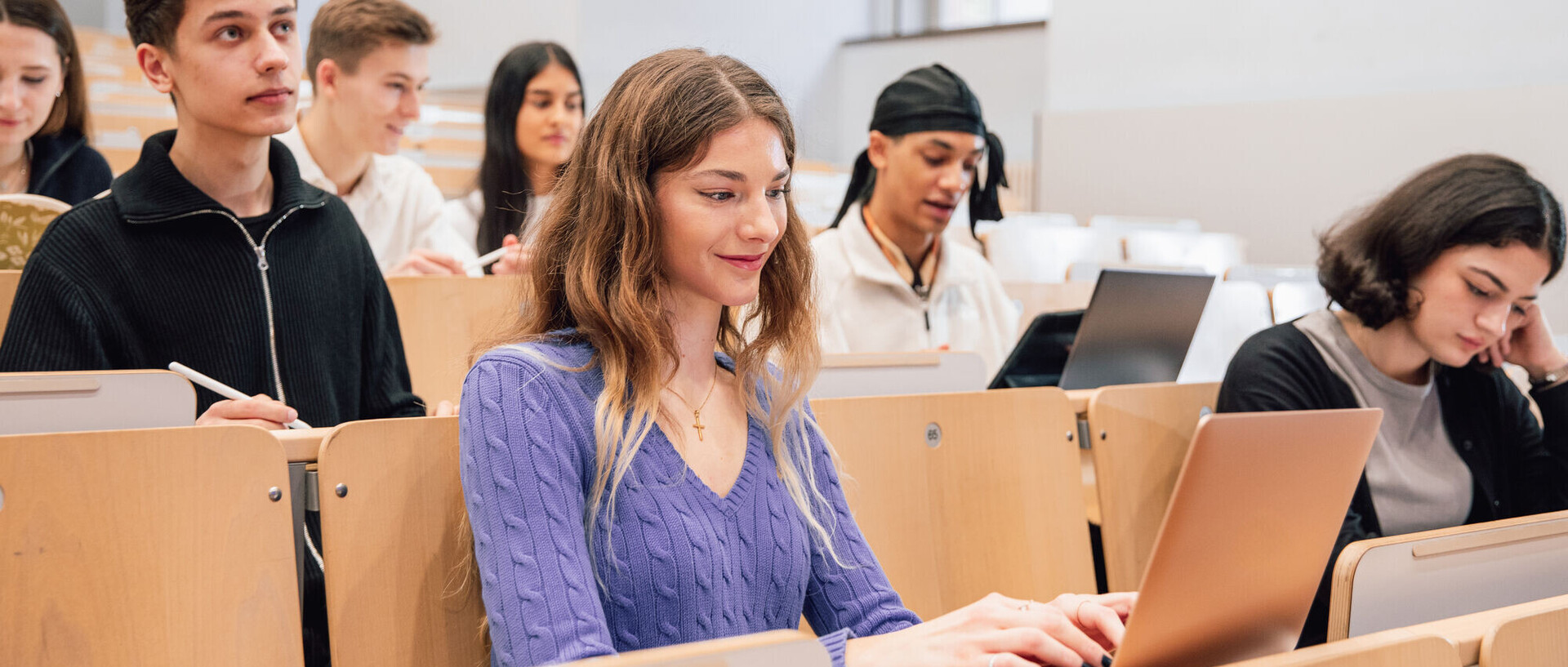 Students sit in a lecture hall. Some have a laptop in front of them, others hold a pen in their hand.