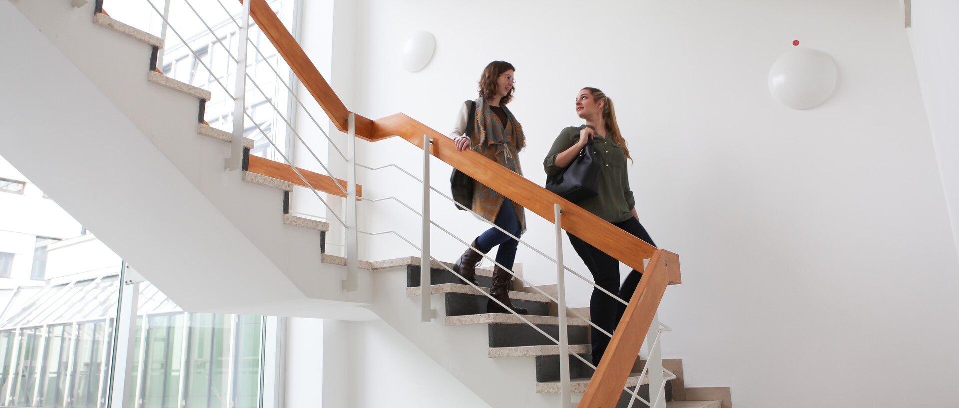 Two female students talk as they walk down the stairs in a white stairwell.